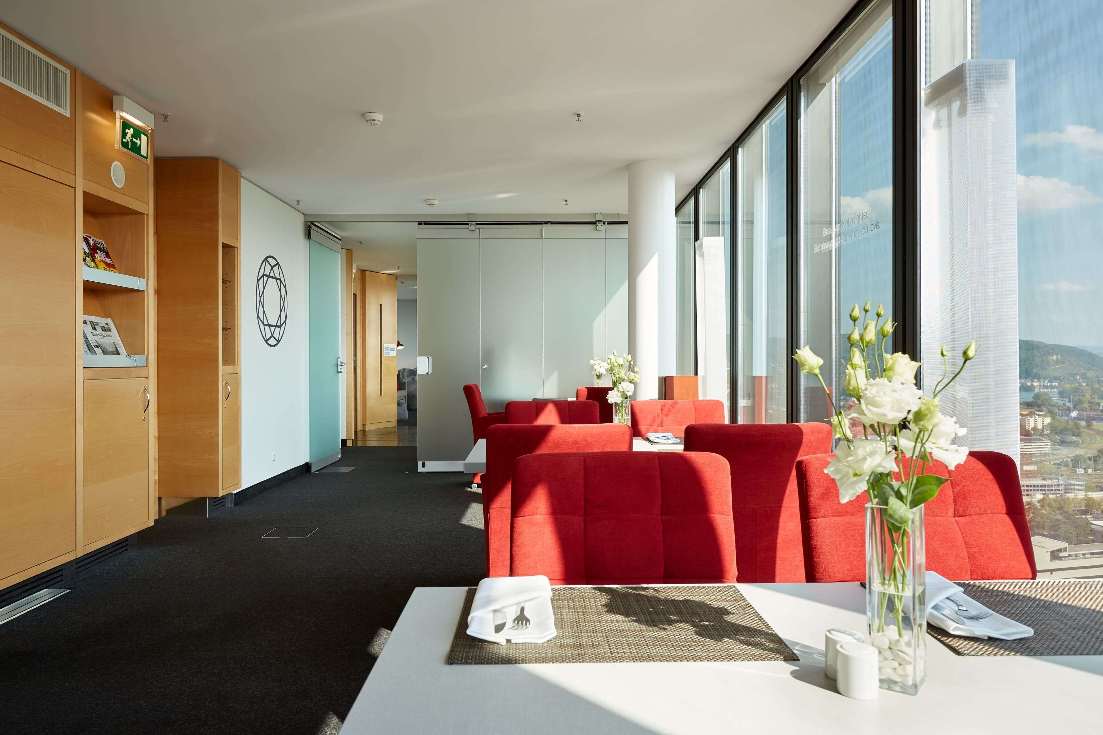 A modern hotel room with a large window, red upholstered seating, a white table, and a wooden cabinet with a green emergency exit sign.