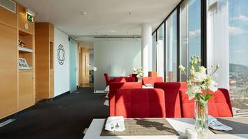 A modern hotel room with a large window, red upholstered seating, a white table, and a wooden cabinet with a green emergency exit sign.