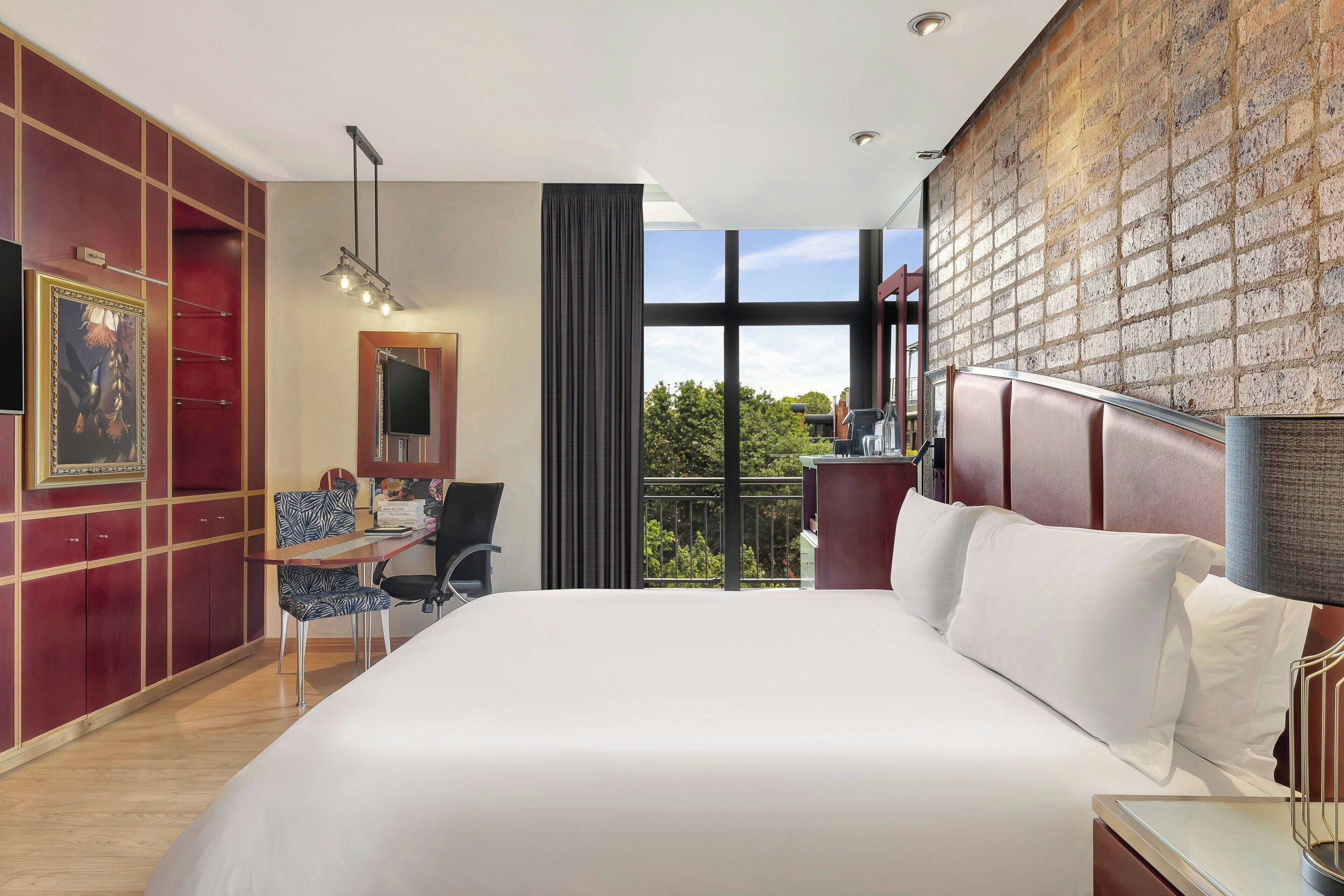 A hotel room with a large bed, a desk, a chair, and a view of the outdoors through large windows.