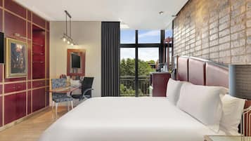 A hotel room with a large bed, a desk, a chair, and a view of the outdoors through large windows.
