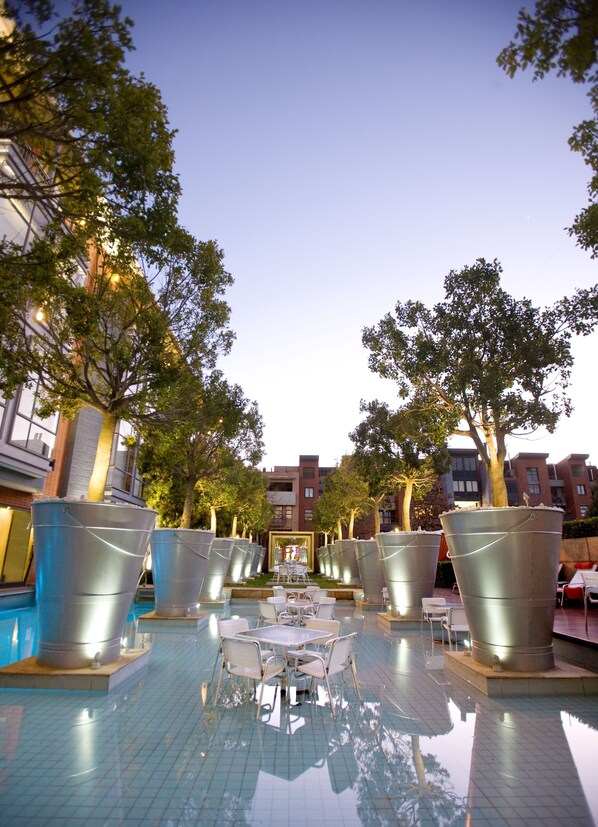Uma área de piscina com vasos de metal, mesas e cadeiras, cercada por árvores e edifícios.