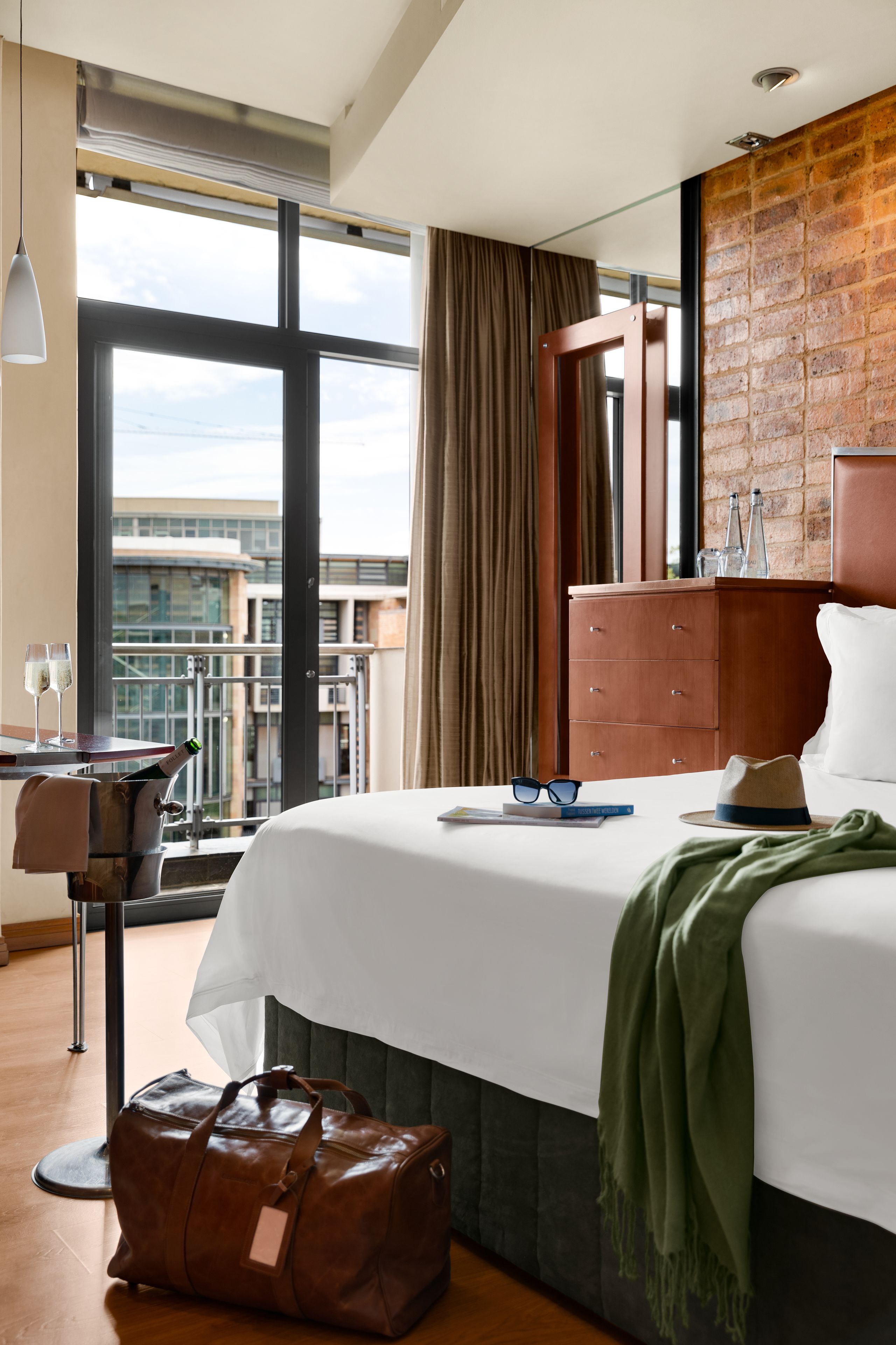 Habitación de hotel con una cama grande, un aparador de madera, una manta verde y vistas de la ciudad a través de amplios ventanales.