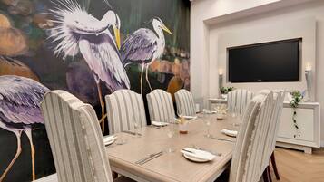 Un comedor con una mesa grande dispuesta para seis personas, rodeada de sillas a rayas, un cuadro de pájaros en la pared y un televisor de pantalla plana montado sobre la mesa.