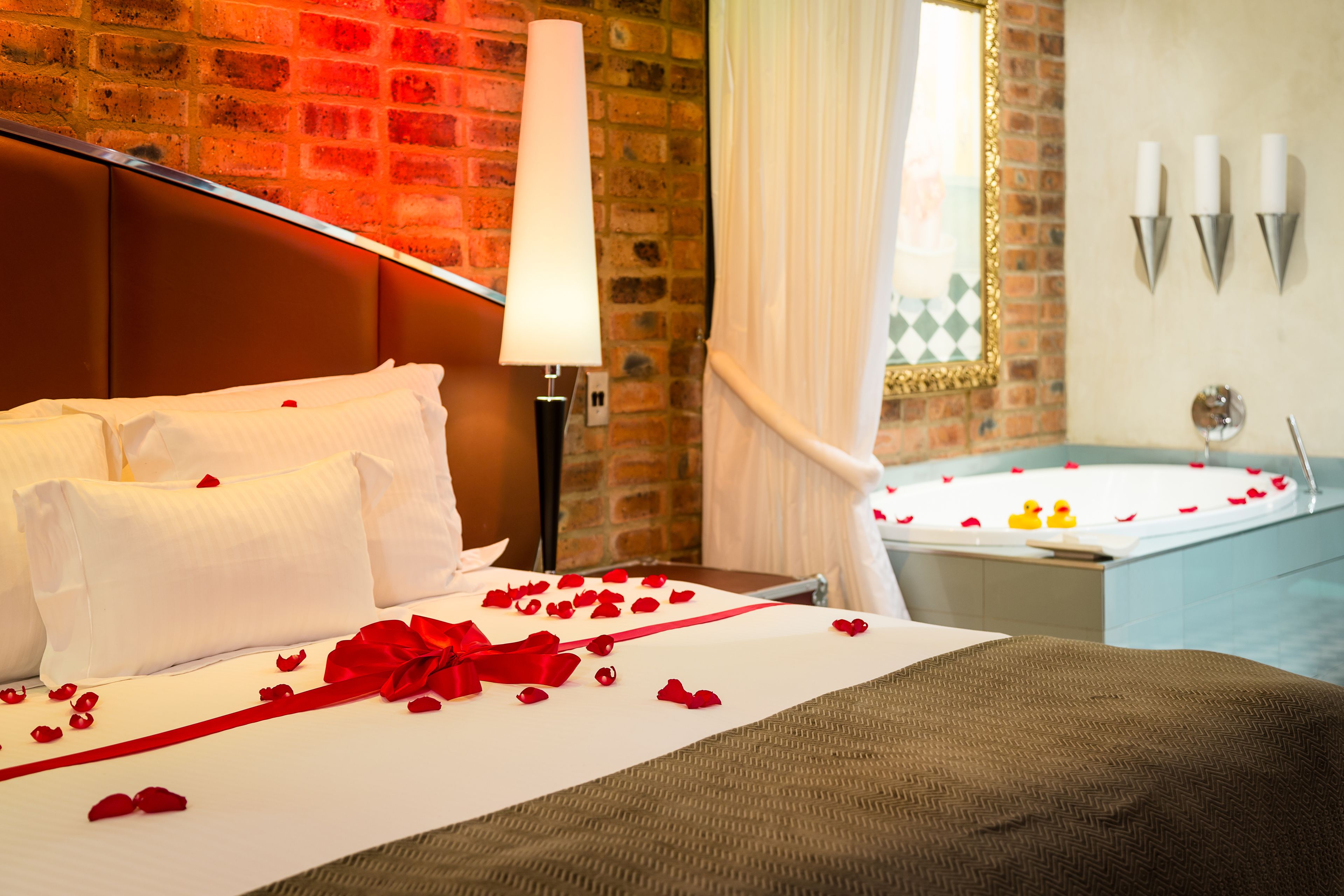 A hotel room with a brick wall, a bed with white linens and red rose petals, a red ribbon, and a bathtub with two rubber ducks.