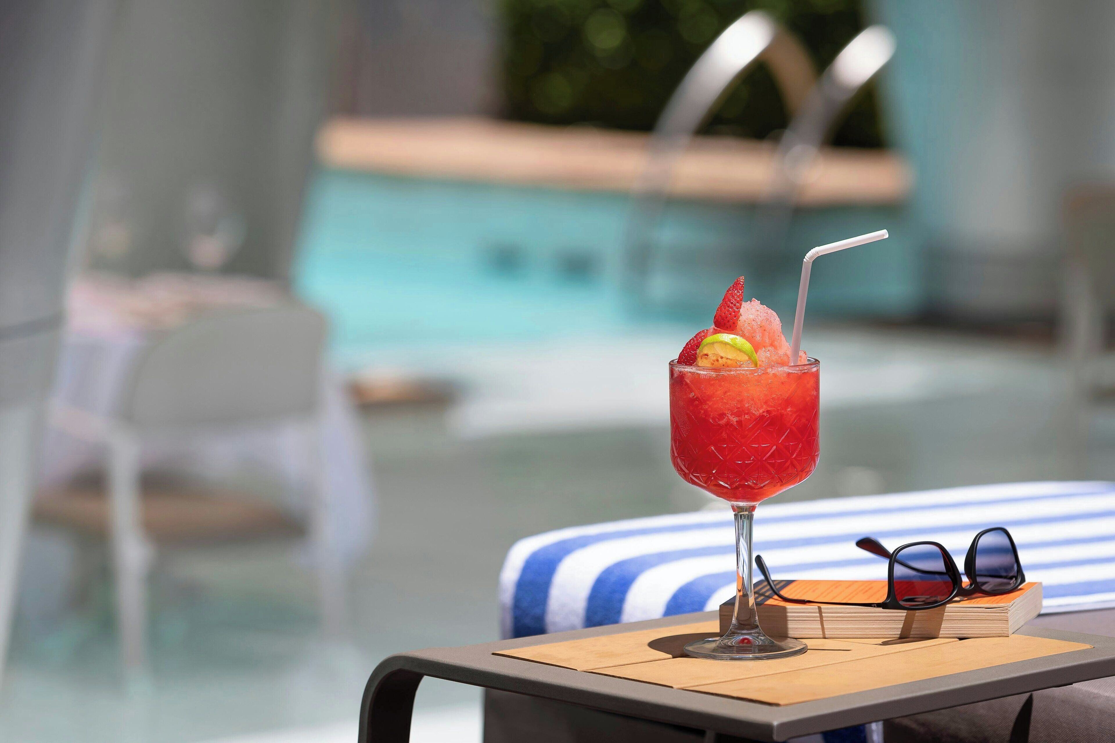 Et glas rød cocktail med sugerør og et jordbær på et bord nær en pool.
