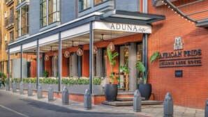 Exterior del Hotel Aduna African Pride Melrose Arch, con entrada cubierta, plantas en macetas grandes y fachada de ladrillo.