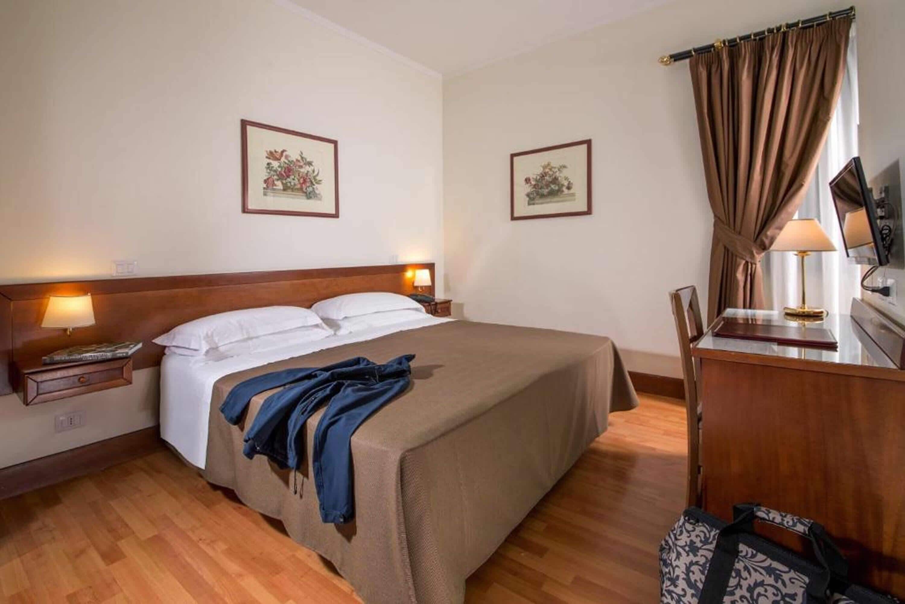 A hotel room with a bed, bedside lamps, a desk with a chair, and two framed pictures on the wall.