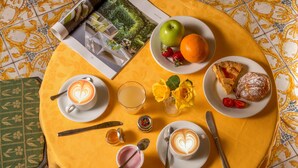 Una mesa con mantel amarillo, dos tazas de café con diseños de corazones en la leche, un plato con una manzana, una naranja, fresas, un pastel, un vaso de jugo, un tarro pequeño de miel y un libro.