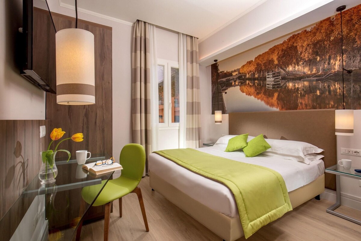 Habitación de hotel con cama, escritorio y silla, un jarrón con flores y un cuadro en la pared.