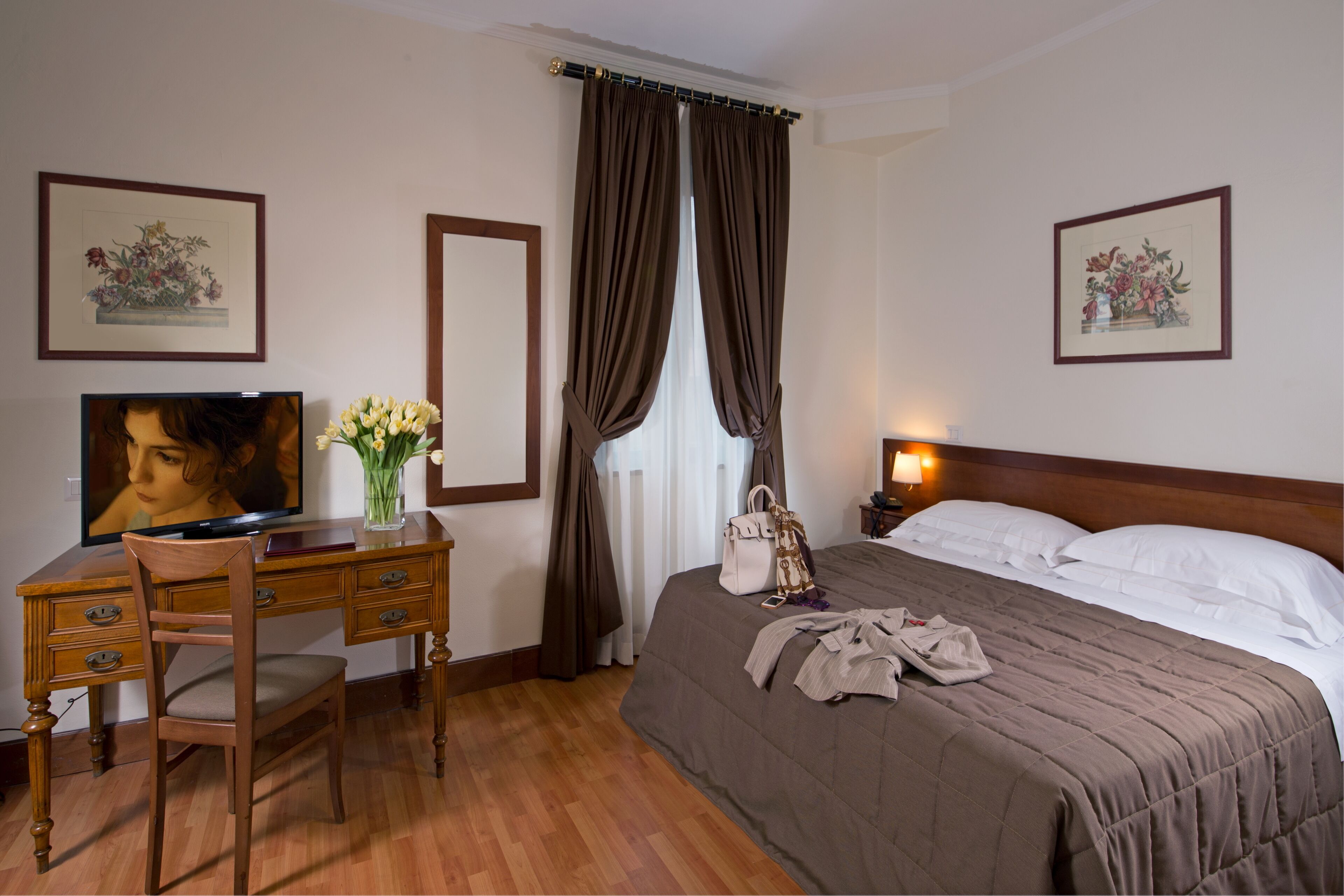 A hotel room with a wooden desk, a chair, a bed with a brown bedspread, a nightstand with a lamp, and two framed floral pictures on the walls.