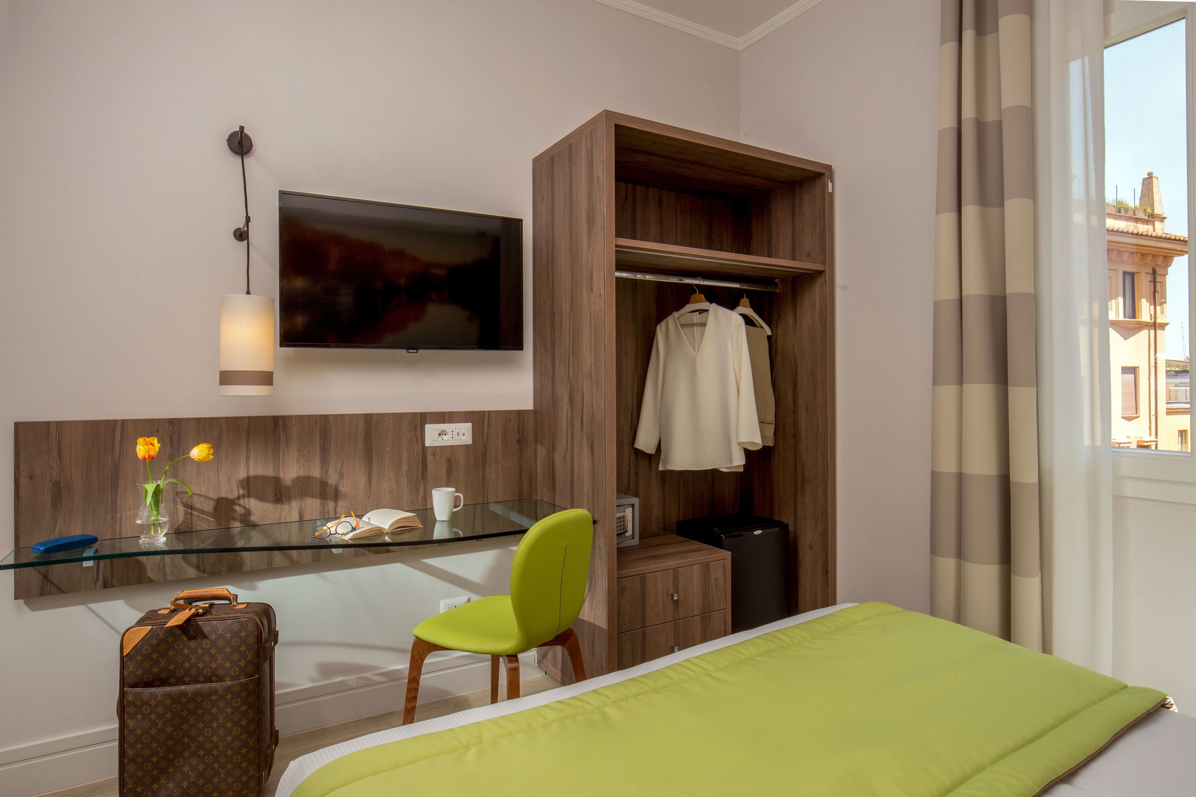 A hotel room with a bed, a desk with a chair, a wardrobe, a TV mounted on the wall, and a window with curtains.