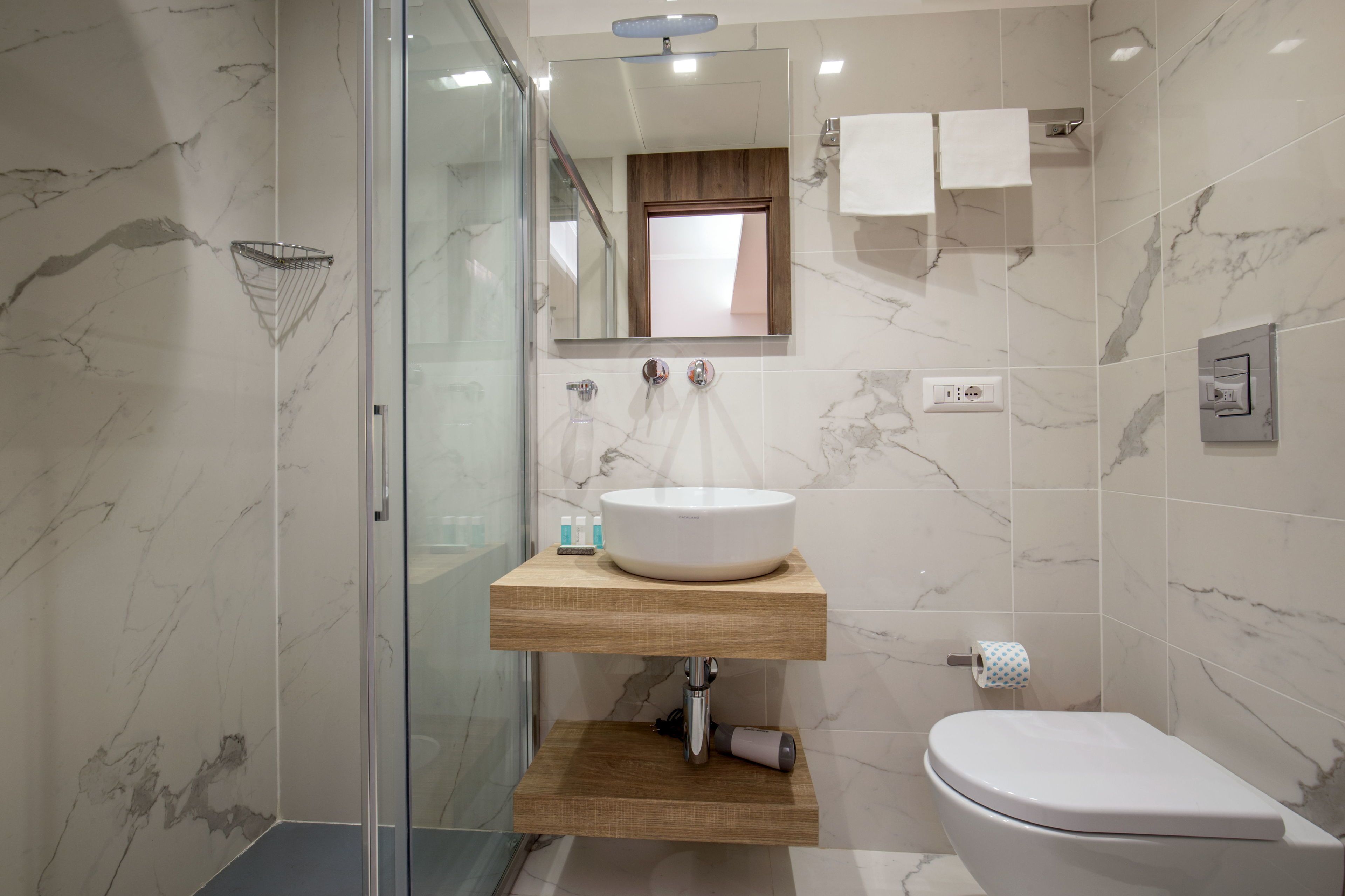 A bathroom with a marble wall, a wooden vanity, a white sink, a toilet, and a glass shower enclosure.
