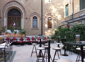 An outdoor café with tables and chairs, potted plants, and a stone building in the background.