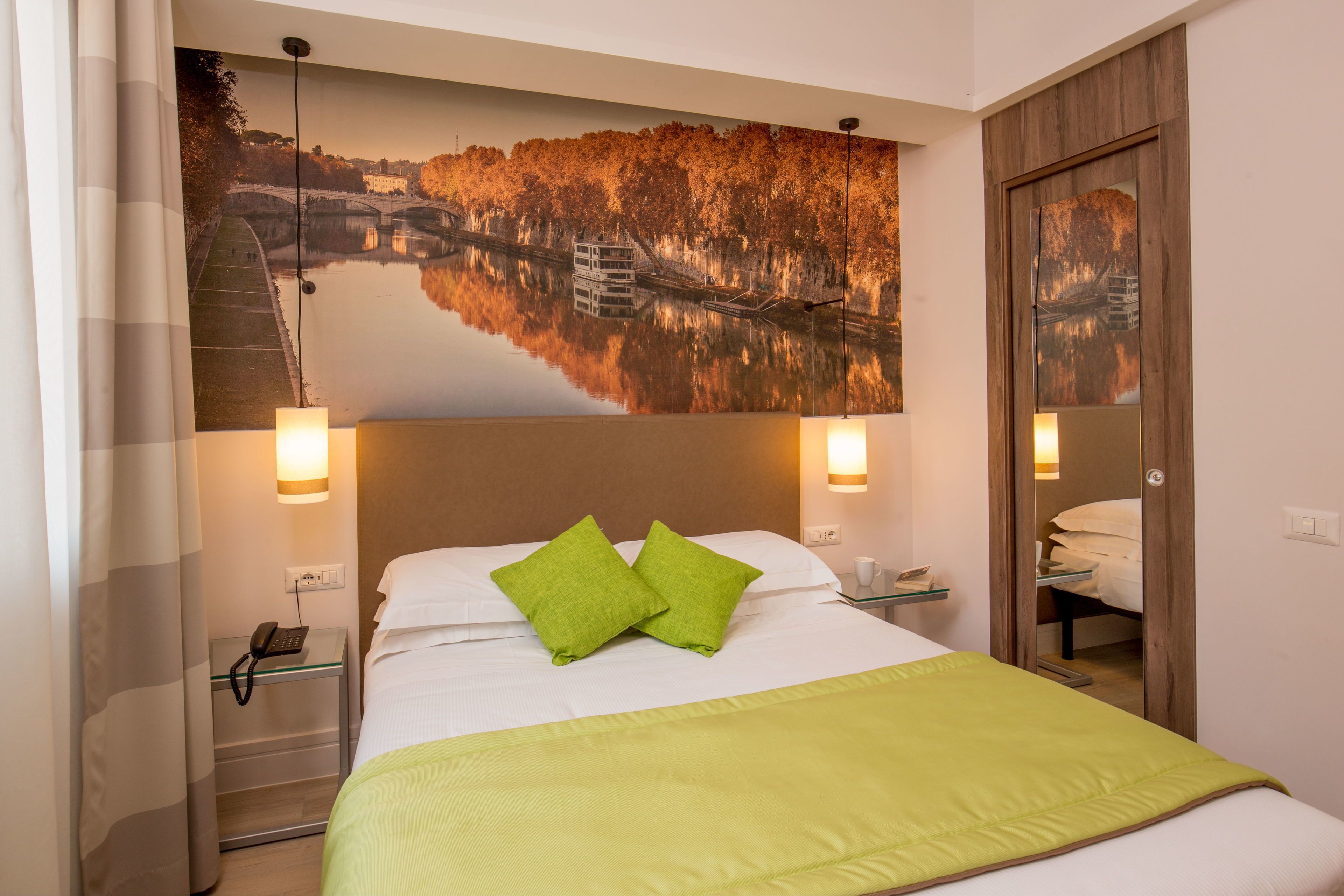 Une chambre d’hôtel avec un lit, des oreillers verts, une table de chevet avec un téléphone et un tableau représentant un paysage fluvial accroché au mur.