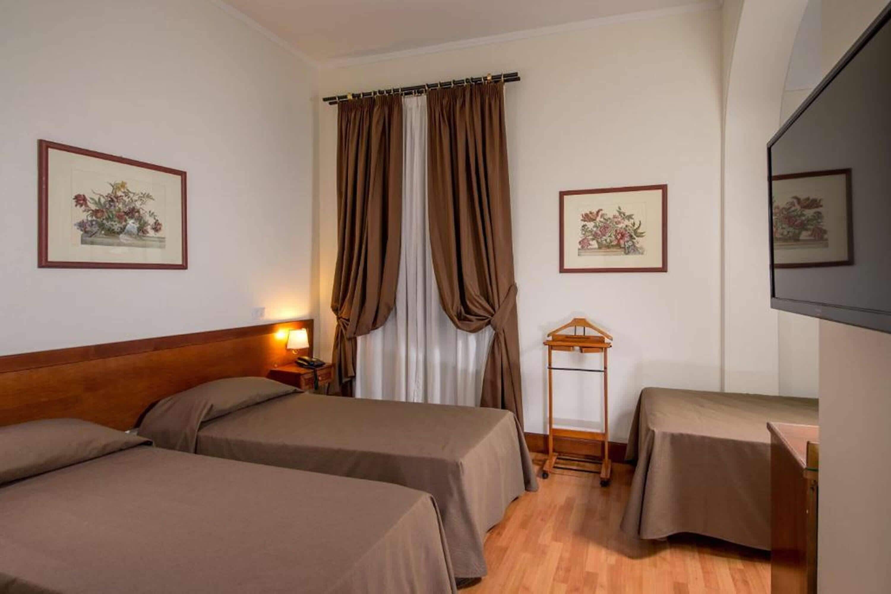 Habitación de hotel con dos camas, cabecera de madera, mesita de noche con lámpara, una pequeña mesa con mantel, y dos cuadros con flores en las paredes.