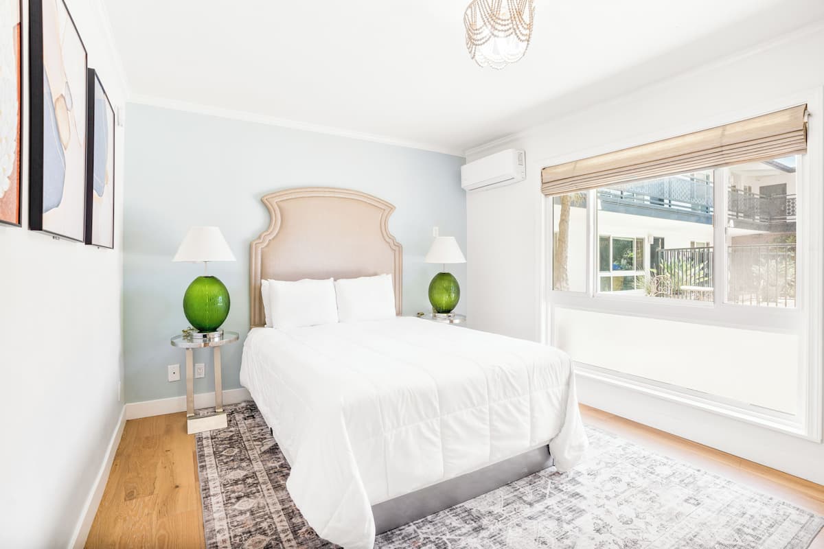 A bedroom with a bed, two bedside lamps, a window with a view, and a patterned rug.