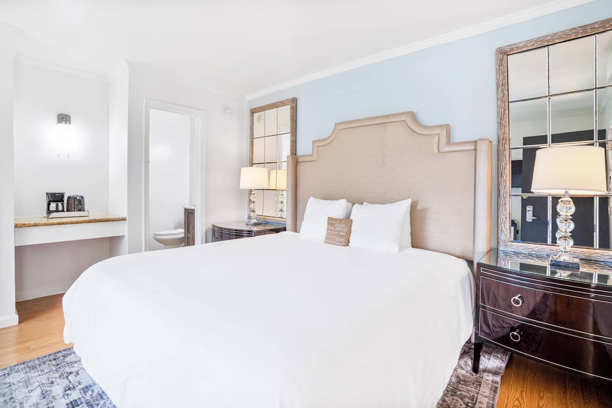 A hotel room with a large bed, a dresser, a nightstand, a mirror, and a small table with a lamp.