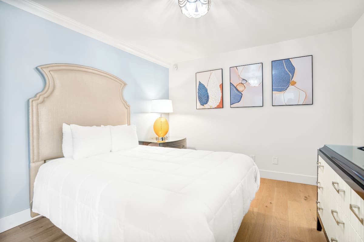 A bedroom with a large bed, a nightstand with a lamp, and three framed artworks on the wall.