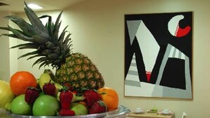 A dining area with a fruit bowl on a table, chairs, a small table with a drink, and a wall with a geometric painting. - Hotel Touring Pisa (Pisa)