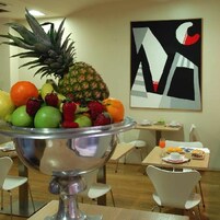 A dining area with a fruit bowl on a table, chairs, a small table with a drink, and a wall with a geometric painting.