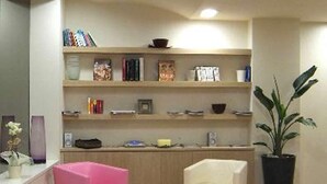 A modern living room with two pink chairs, a white chair, and a bookshelf filled with books and decorative items. - Hotel Touring Pisa (Pisa)