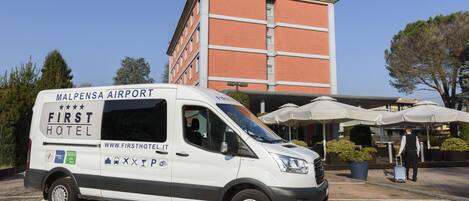 Una camioneta blanca con la leyenda "Malpensa Airport" y "First Hotel" estacionada frente a un edificio de hotel.