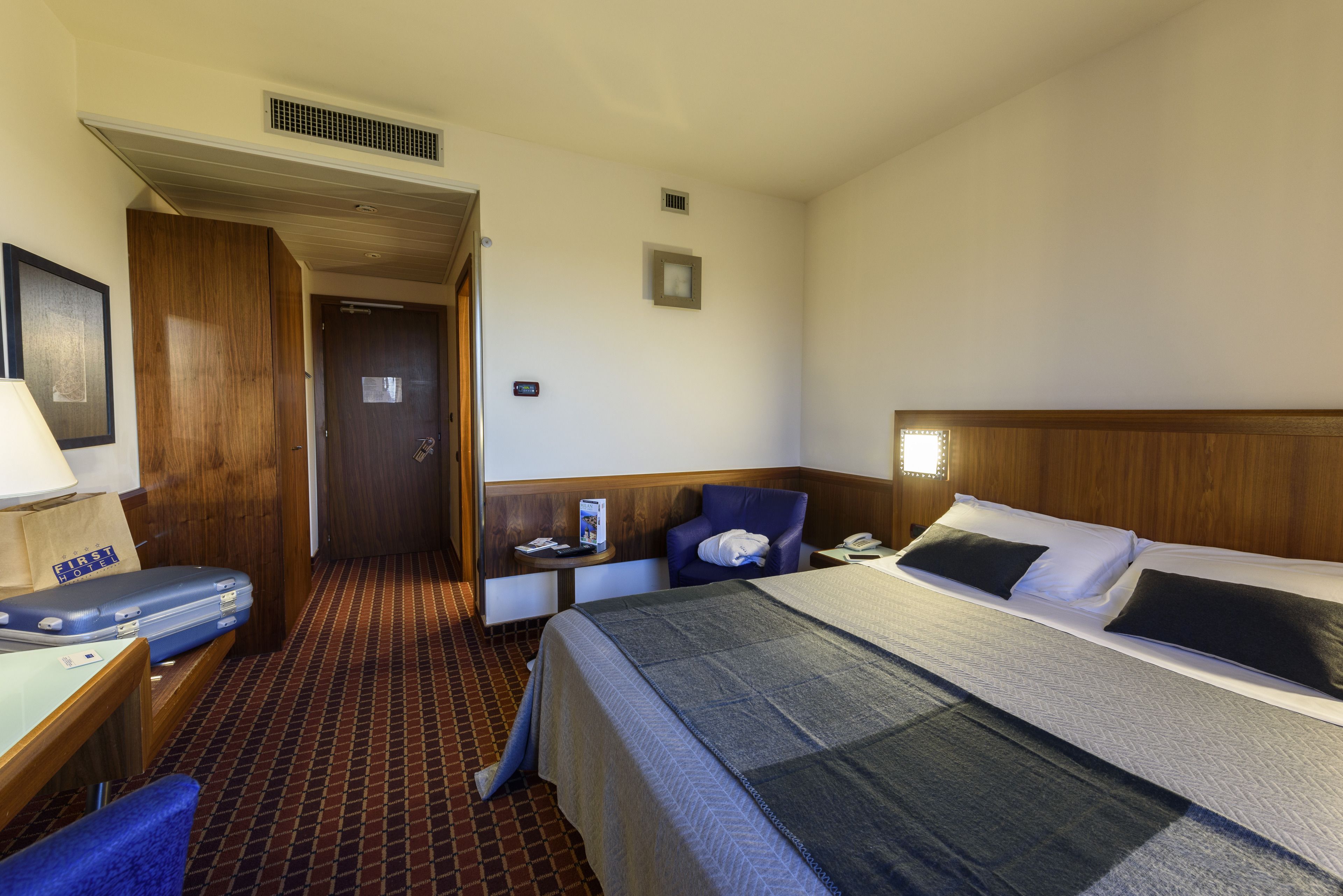 A hotel room with a bed, a desk with a suitcase, a chair, and a small table with a lamp.