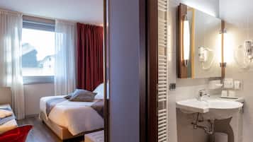 Habitació d'hotel amb un llit, una taula de nit, una cadira vermella, una finestra amb cortines i un bany amb un lavabo i un mirall.