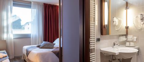 A hotel room with a bed, a bedside table, a red chair, a window with curtains, and a bathroom with a sink and mirror.