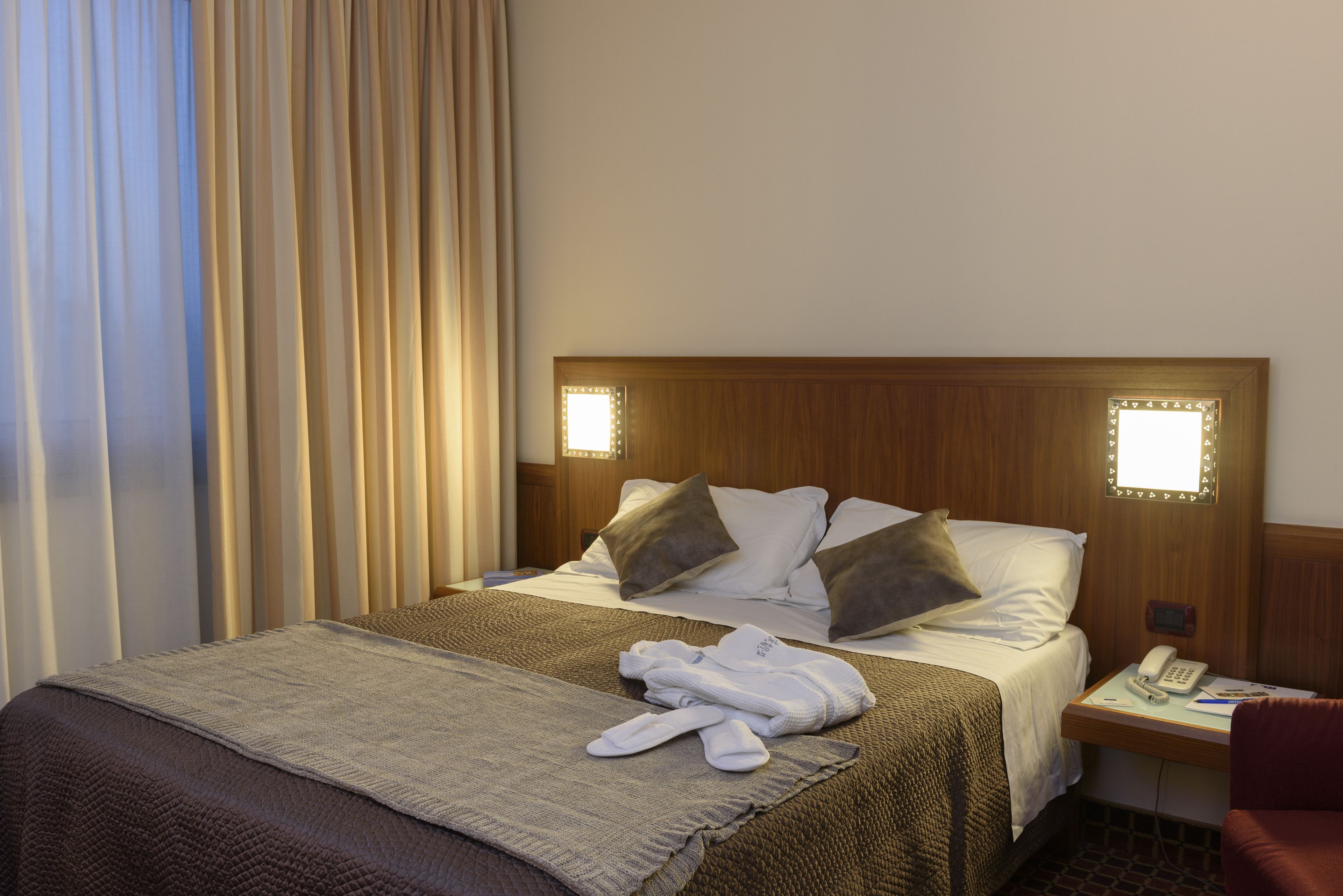 A hotel room with a neatly made bed, a bedside table with a phone and a lamp, and a red chair.