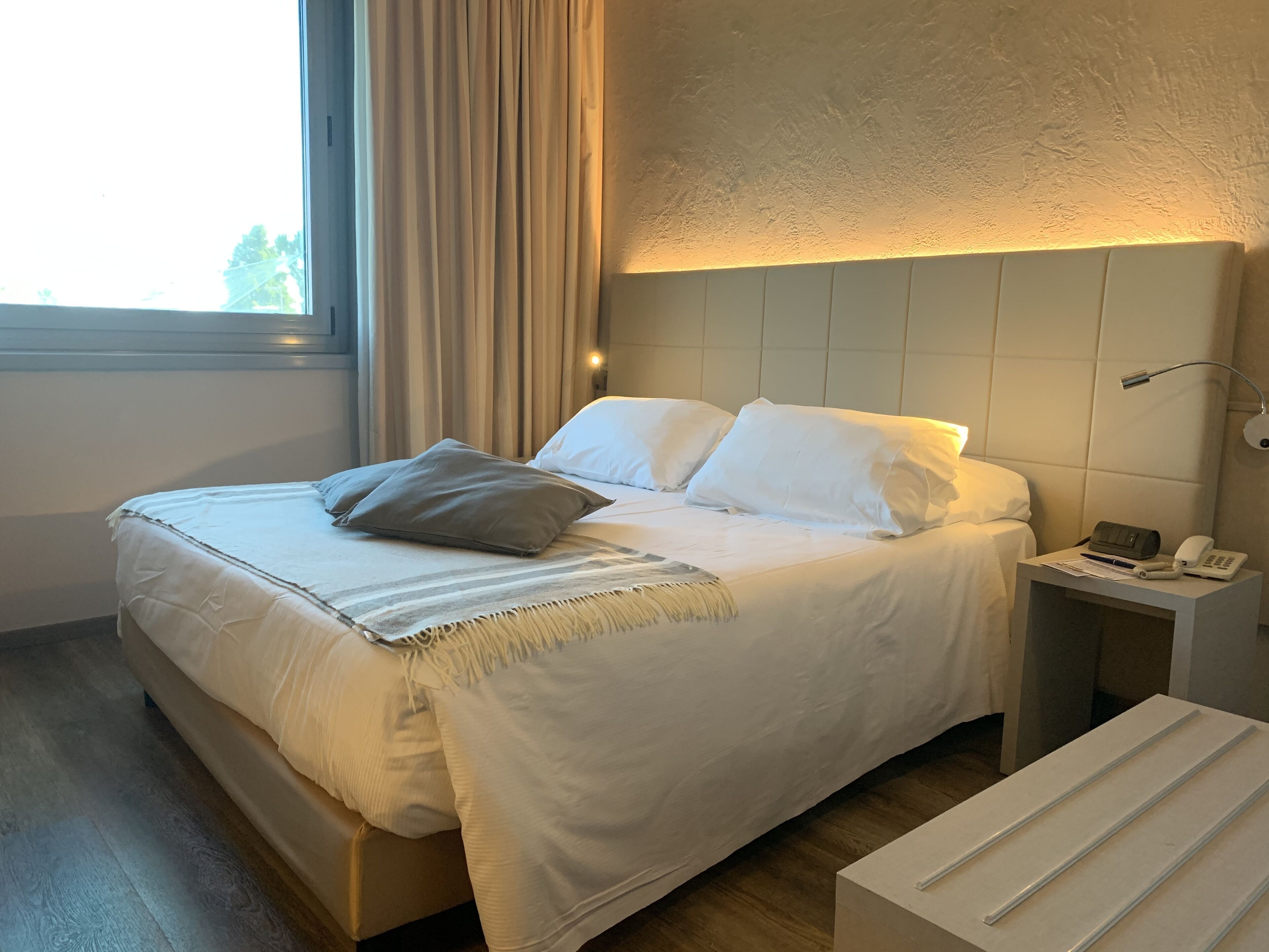 Un letto rifatto con lenzuola bianche e un solo cuscino marrone, un comodino con una lampada e un telefono, e una finestra con tende.