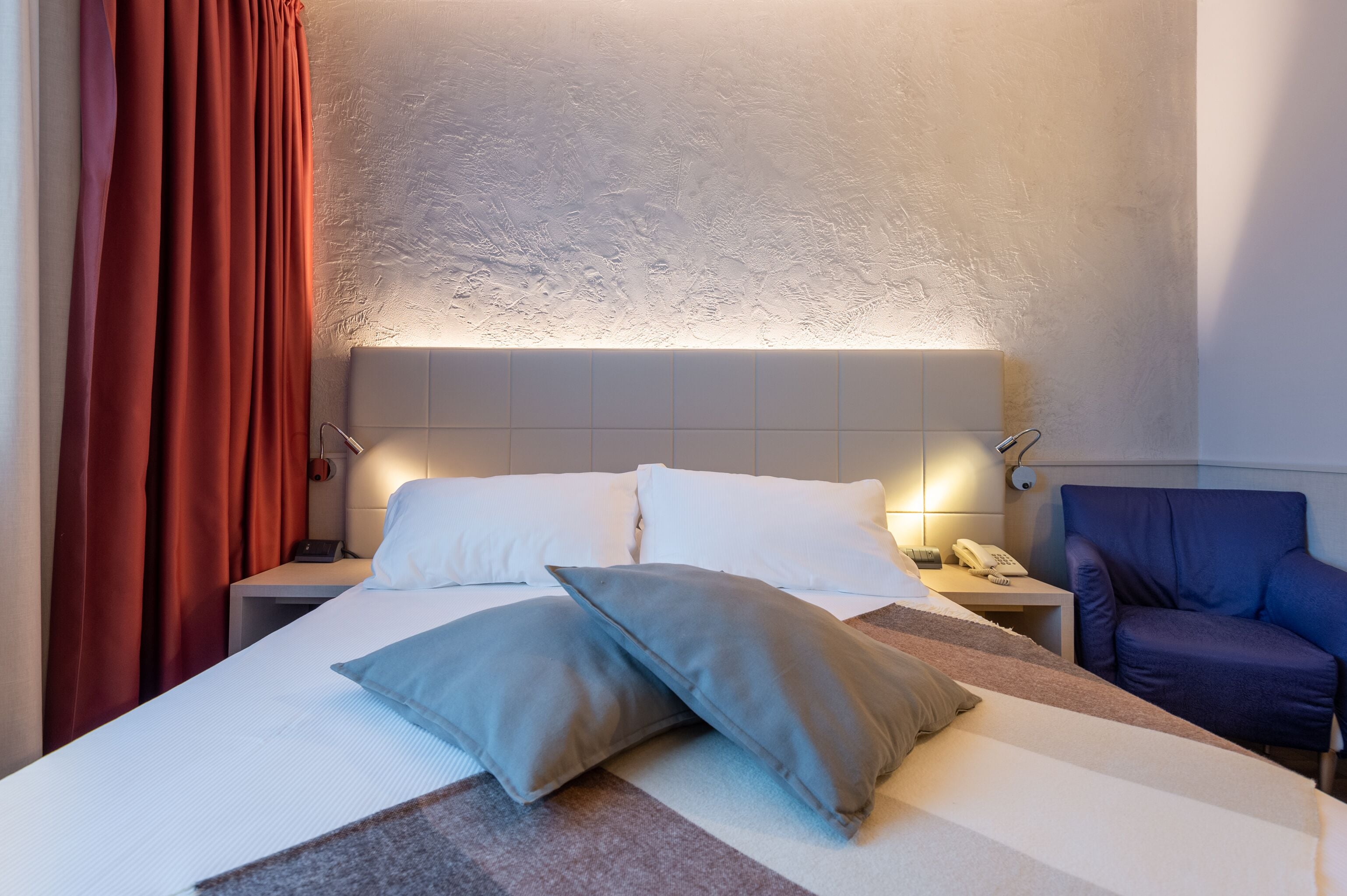 Une chambre d’hôtel avec un grand lit, deux oreillers, une table de chevet avec un téléphone, une chaise et un mur à texture.