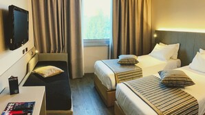 A hotel room with two single beds, a desk with a chair, a flat-screen TV mounted on the wall, and a window with curtains.