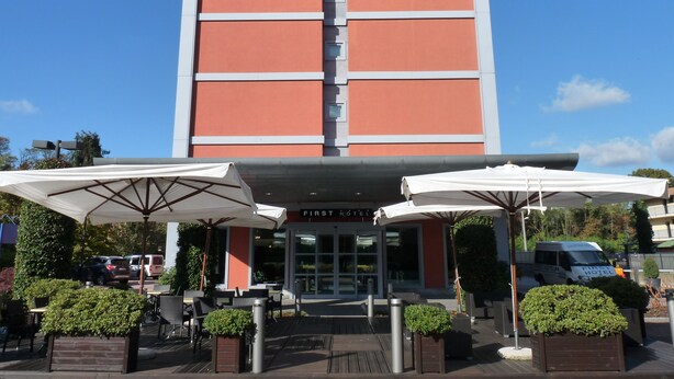 A hotel entrance with a sign reading "FIRST HOTEL". There are outdoor umbrellas and seating areas with planters.