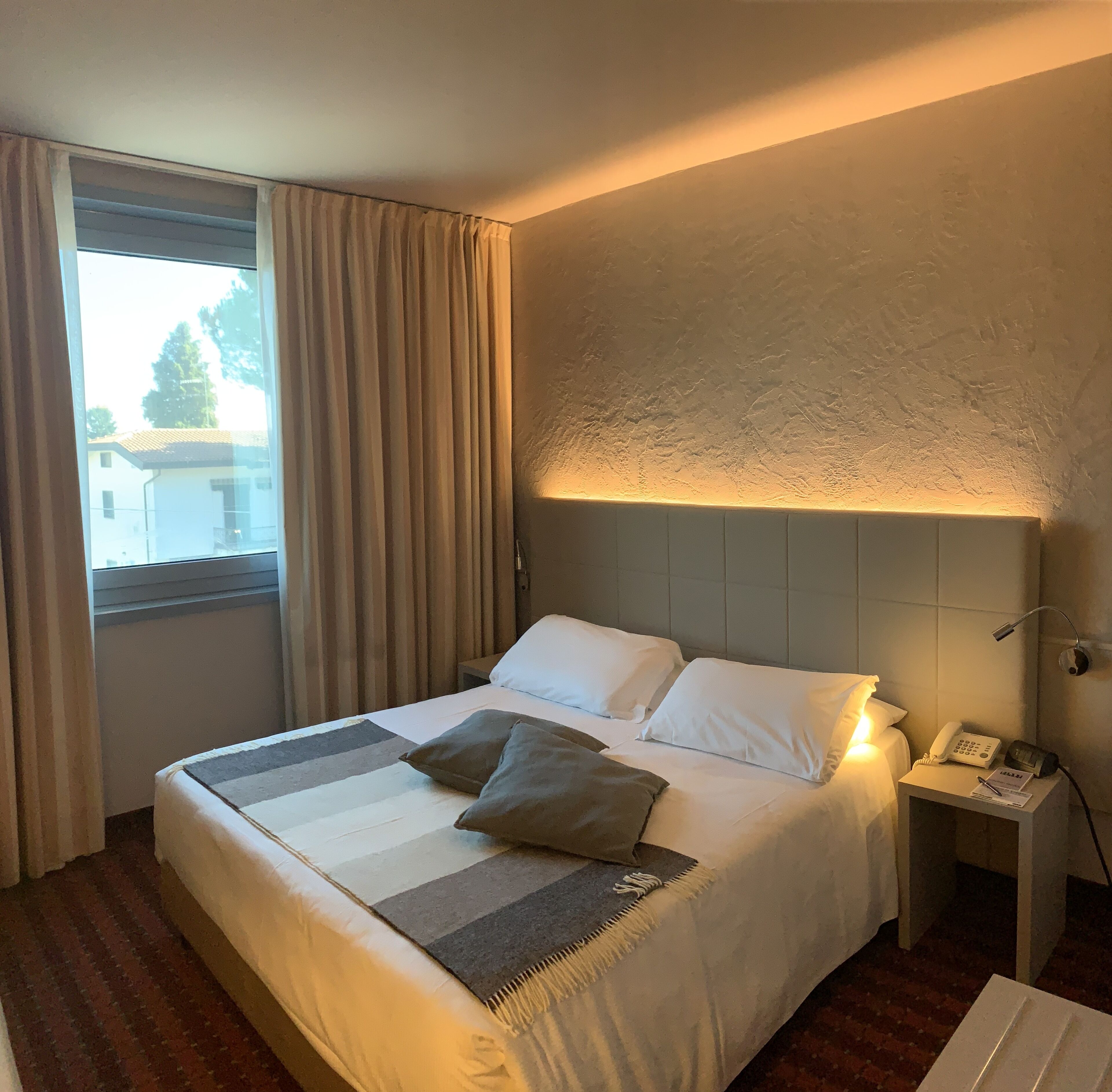 A hotel room with a bed, a nightstand with a phone, and a window with curtains.