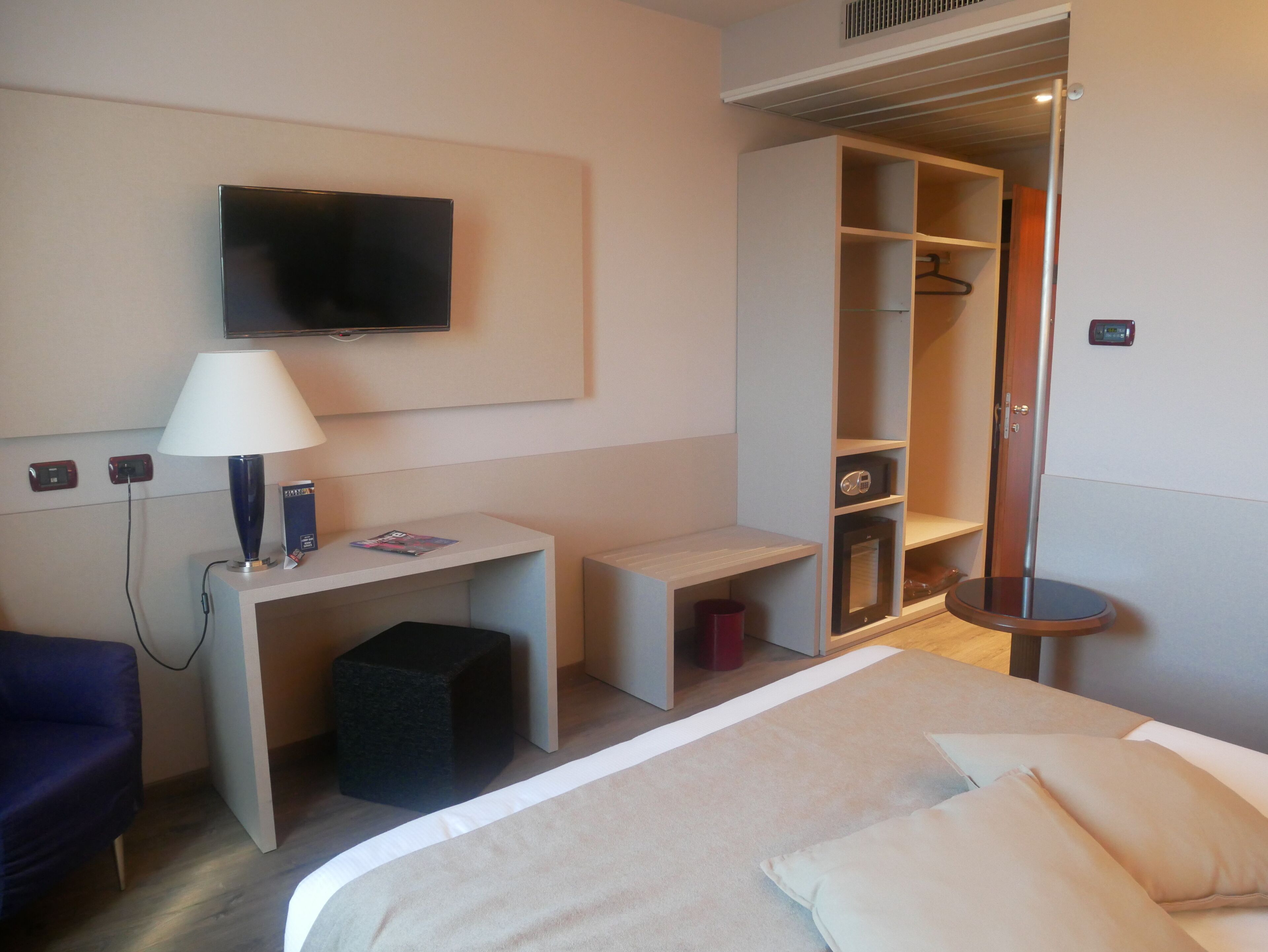 Ein Hotelzimmer mit einem Bett, einem Schreibtisch mit Lampe, einem an der Wand montierten Fernseher und einem begehbaren Kleiderschrank.