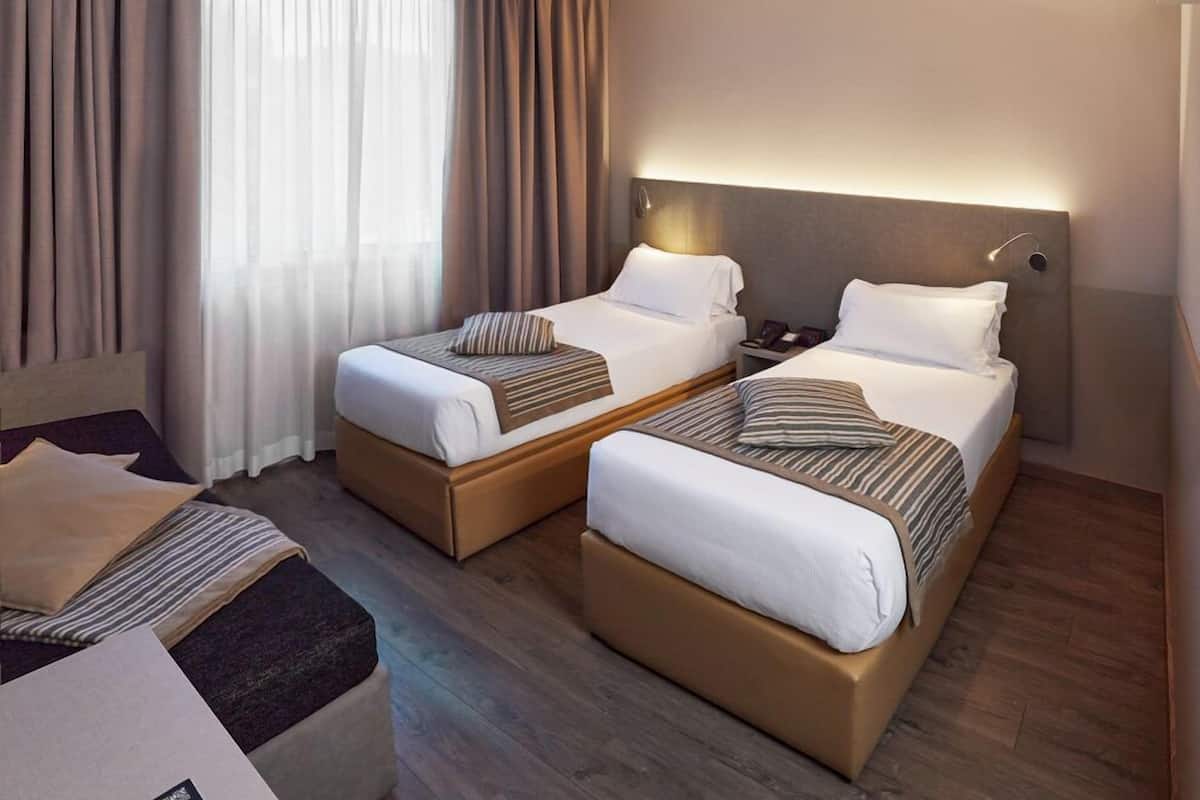 A hotel room with two single beds, a sofa, a desk, and a large window with curtains.