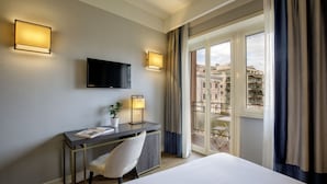 Habitación de hotel con cama, escritorio con silla, televisor de pantalla plana, dos lámparas de pared y balcón con vistas a edificios.