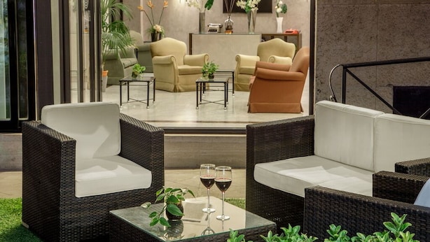 Un espacio exterior moderno con mobiliario de mimbre, una mesa de vidrio con copas de vino y vistas al interior, que incluye un sofá y plantas decorativas.