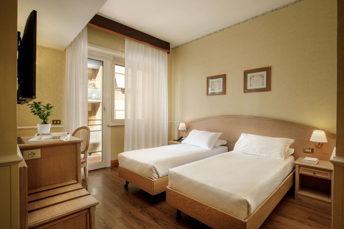 Habitación de hotel con dos camas individuales, un escritorio, una silla, un televisor y una ventana con cortinas.