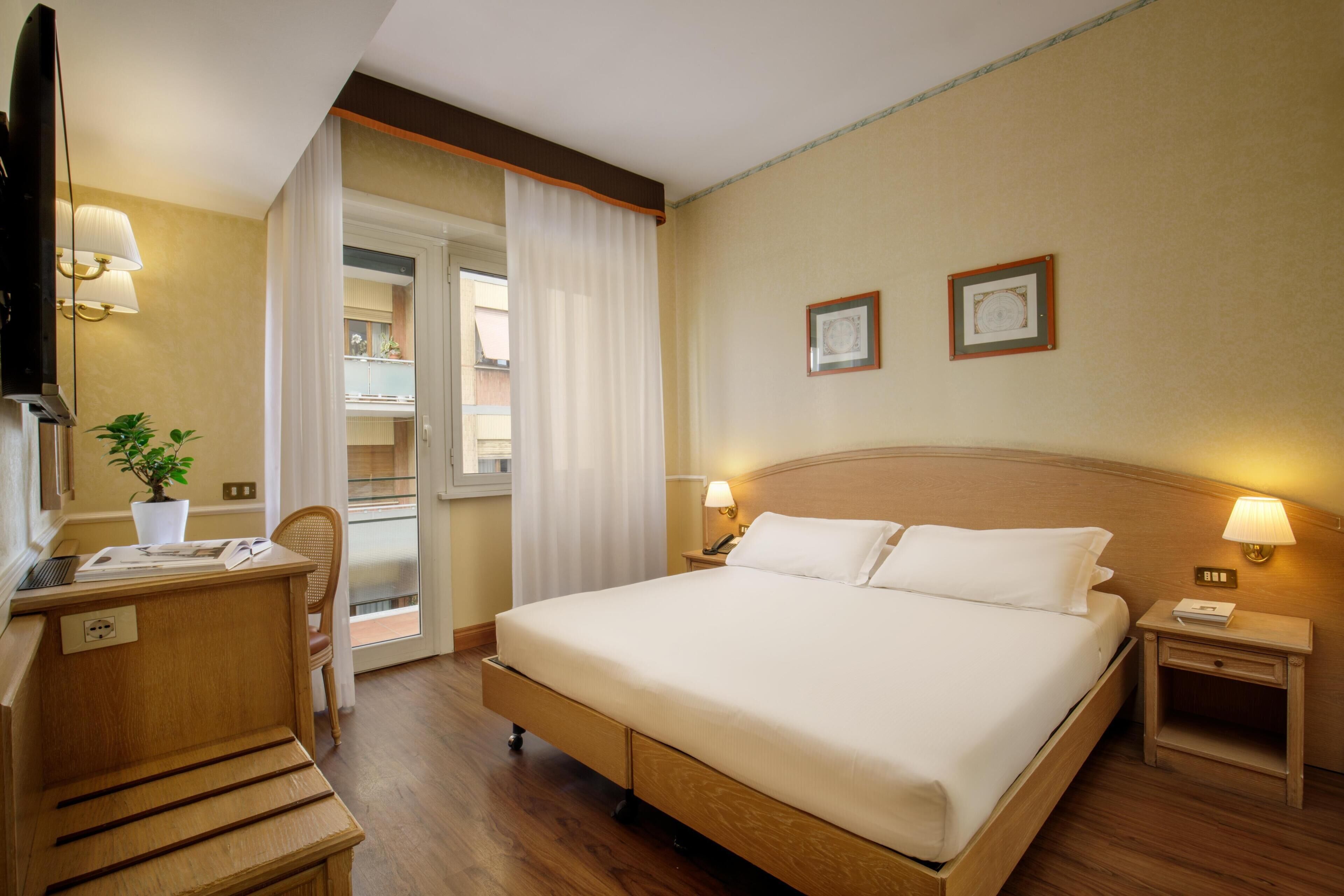 A hotel room with a bed, bedside tables, a desk, a chair, a TV, and a window with curtains.