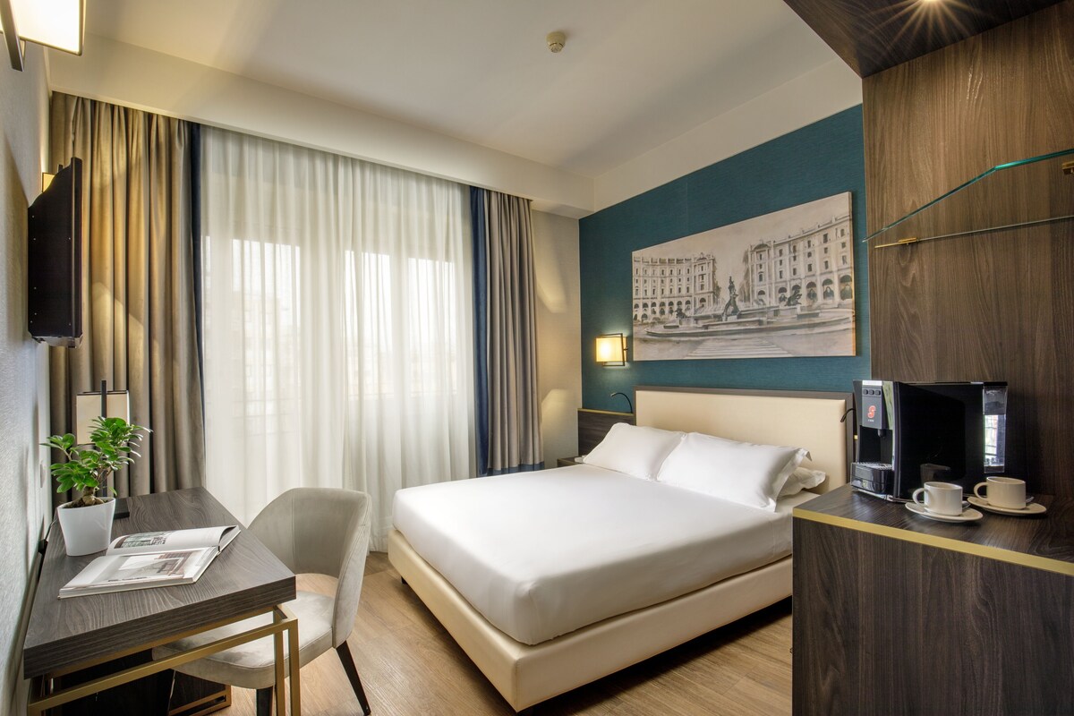 Habitación de hotel con cama, escritorio, silla, cafetera y un cuadro en la pared.