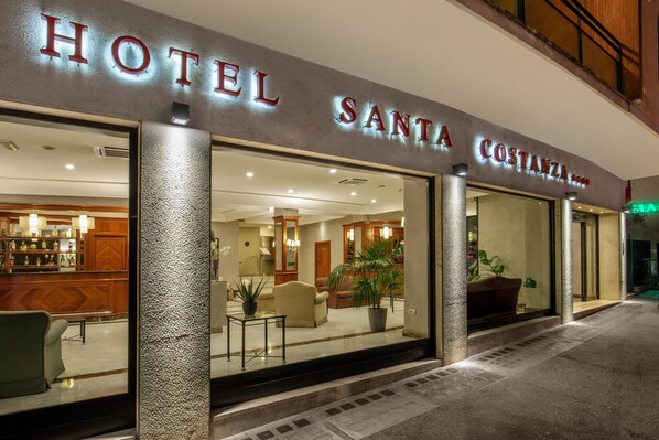 Entrada de un hotel con una zona de bar visible en el interior, una recepción y un área de descanso con sillas y una mesa.