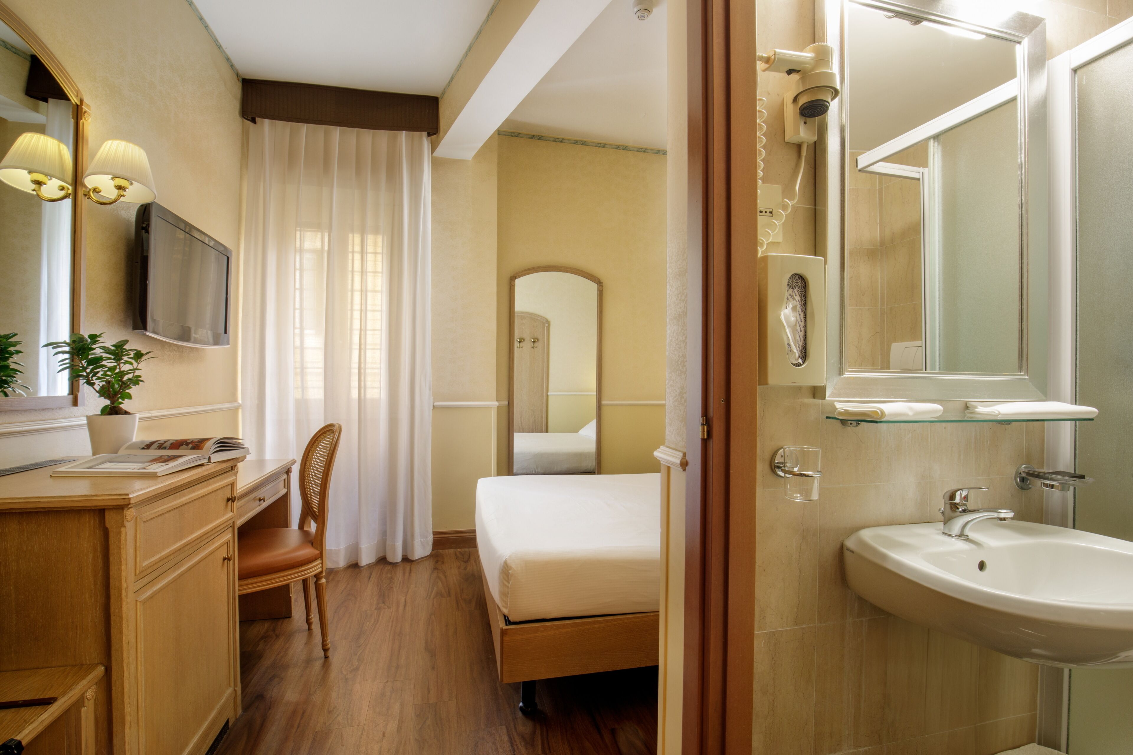 A hotel room with a wooden desk, chair, bed, and a bathroom with a sink and mirror.