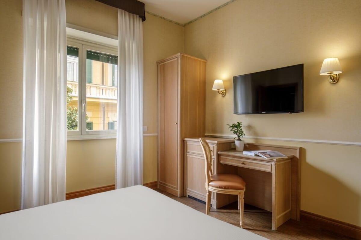 Habitación de hotel con cama, escritorio y silla, televisor y ventana con cortinas.