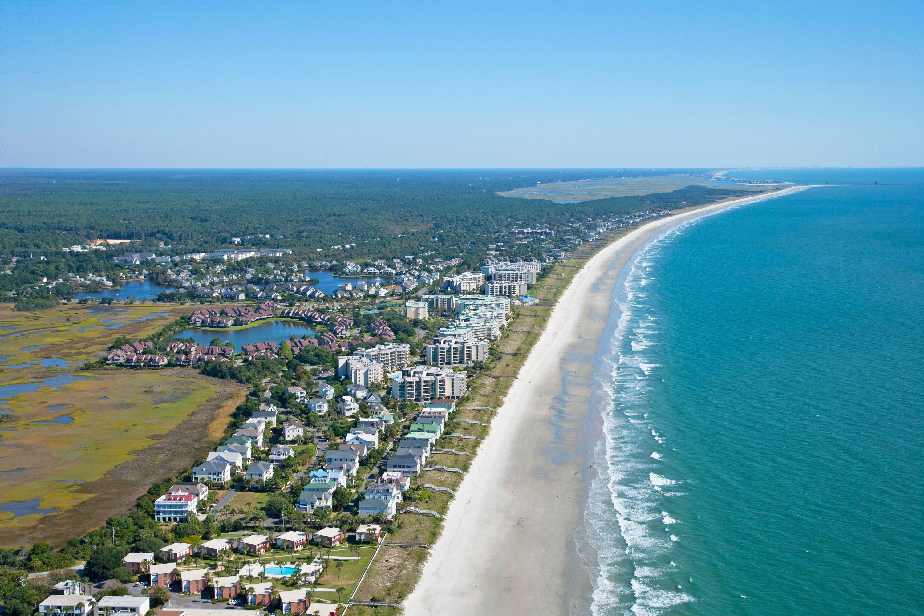 An aerial view of a coastal area with a long sandy beach, a residential neighborhood, and a large body of water.