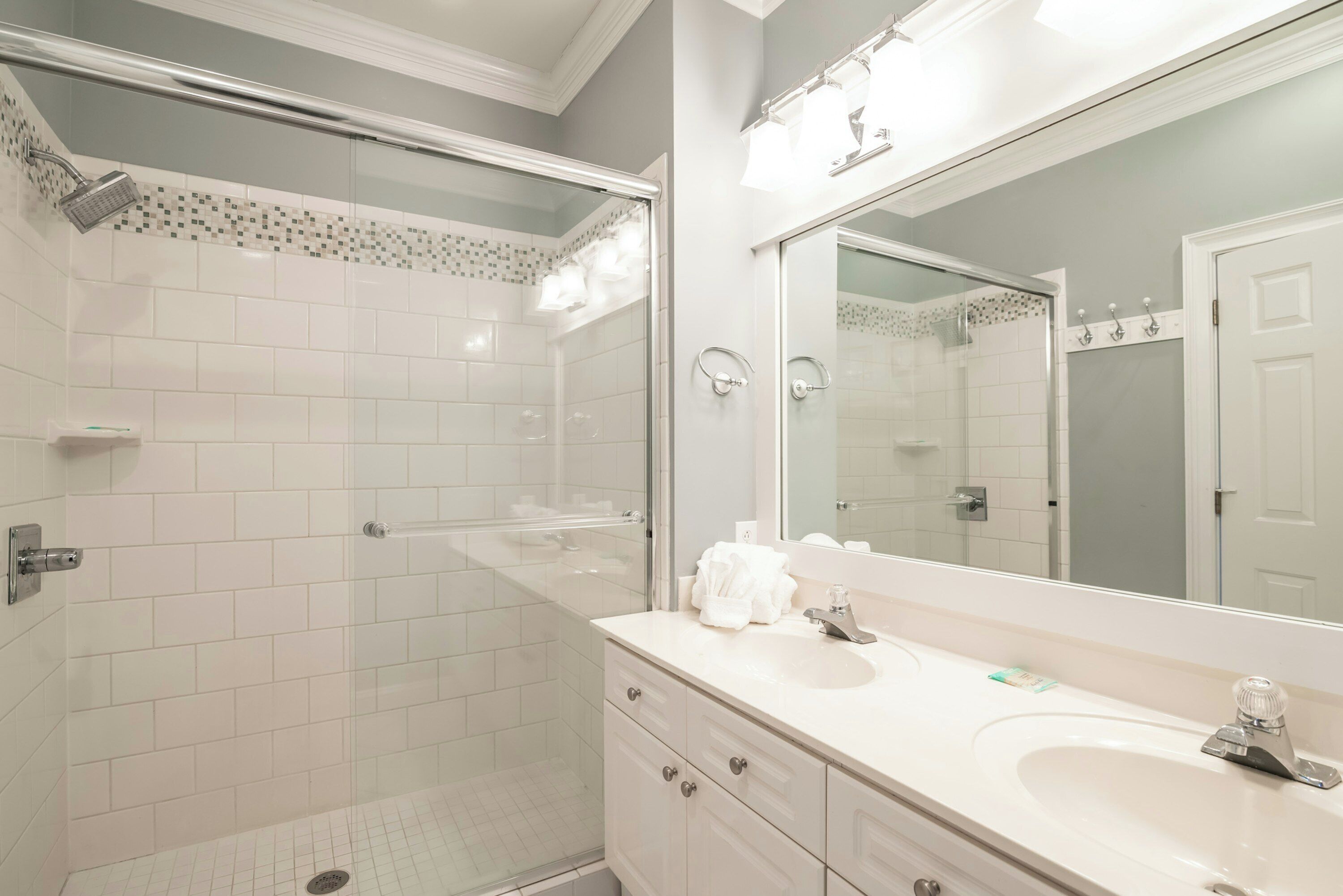 Une salle de bain moderne avec une douche à parois de verre, des murs recouverts de carreaux blancs, un grand miroir et un lavabo blanc.