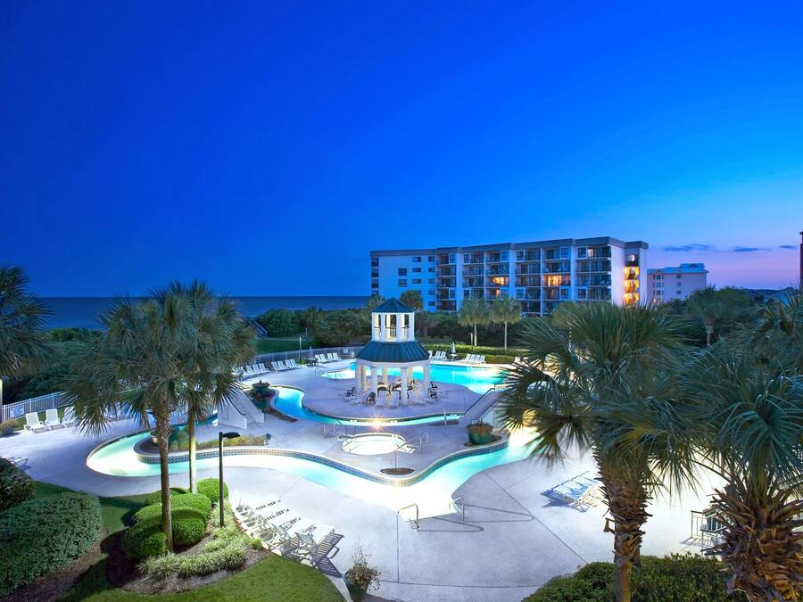 Un espace piscine d’un complexe hôtelier, agrémenté d’un kiosque, de palmiers et offrant une vue sur l’océan au crépuscule.
