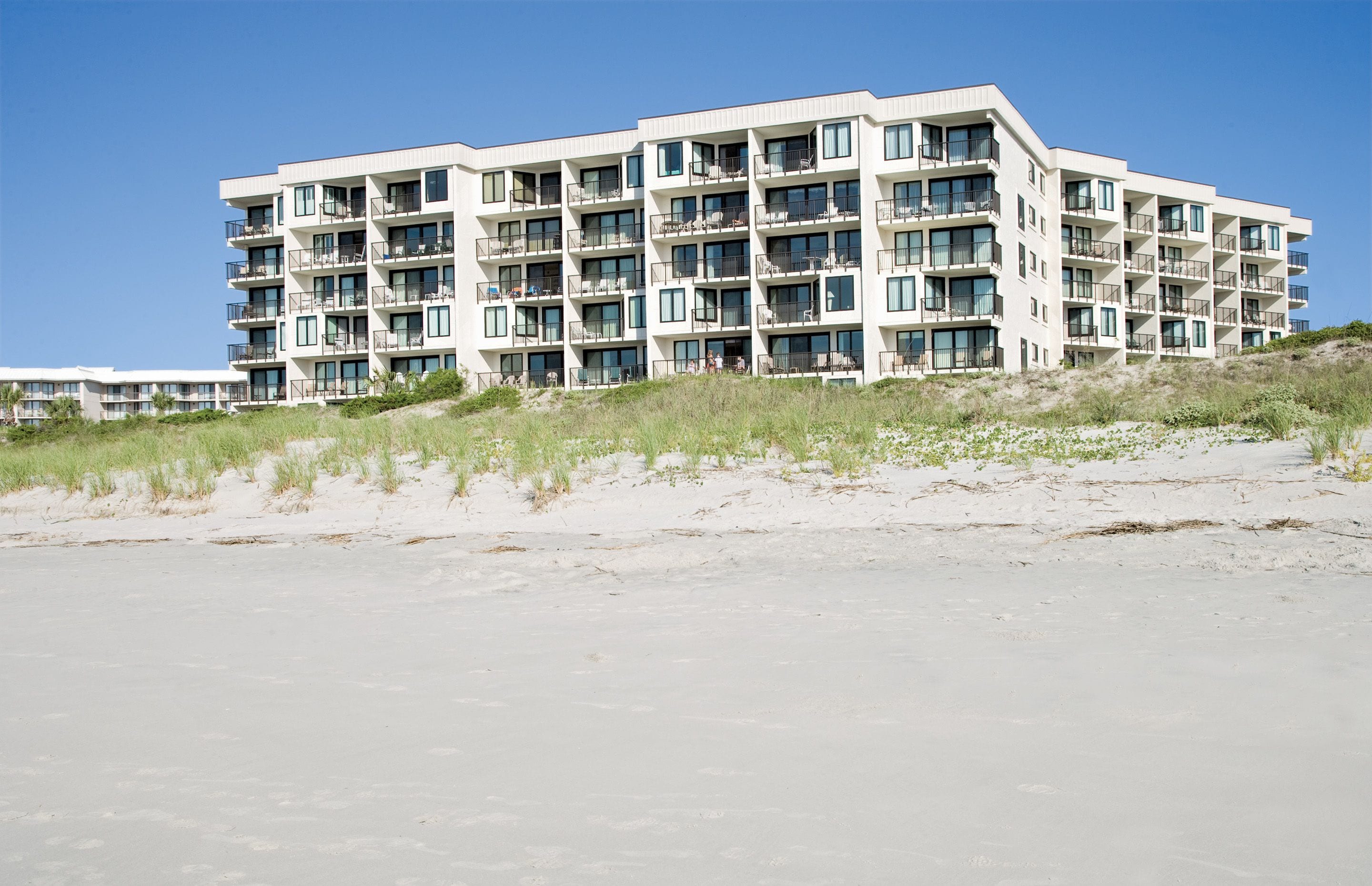 A multi-story hotel with balconies, located on a beach with sand dunes in the foreground.