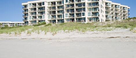 Een hotel met meerdere verdiepingen en balkons, gelegen aan een strand met zandduinen op de voorgrond.